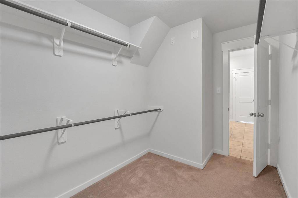 Primary bedroom walk-in closet with double hanging rods, neutral carpeting, and easy access to the utility room with tiled flooring. Perfect for ample storage and organization.