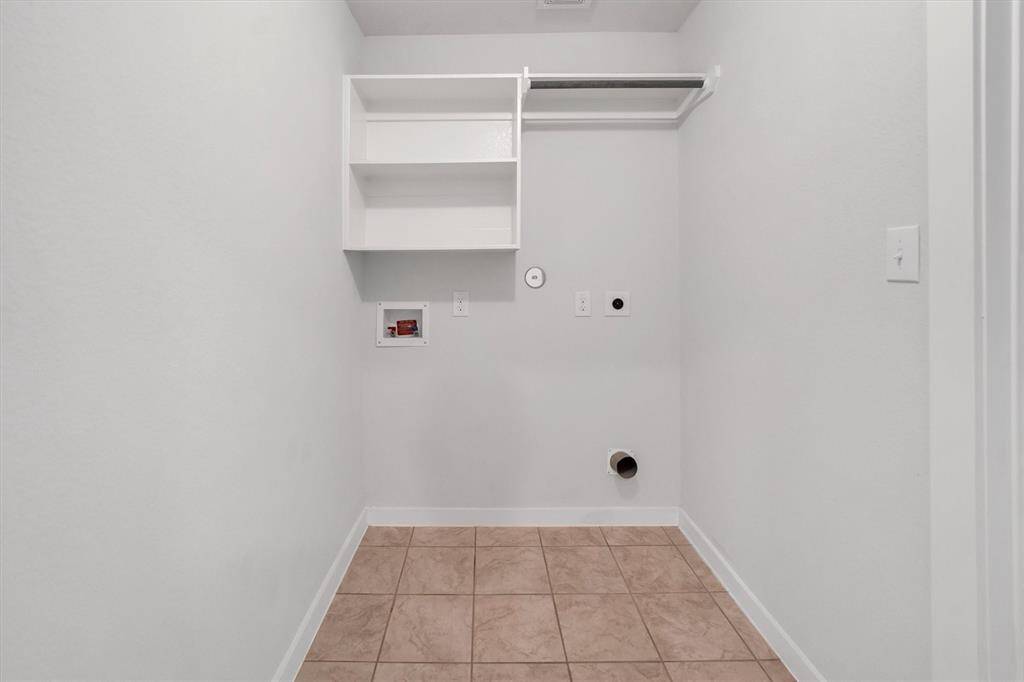 This photo shows a clean, tiled laundry room with hookups for a washer and dryer. It features a white shelf and a hanging rod for additional storage.