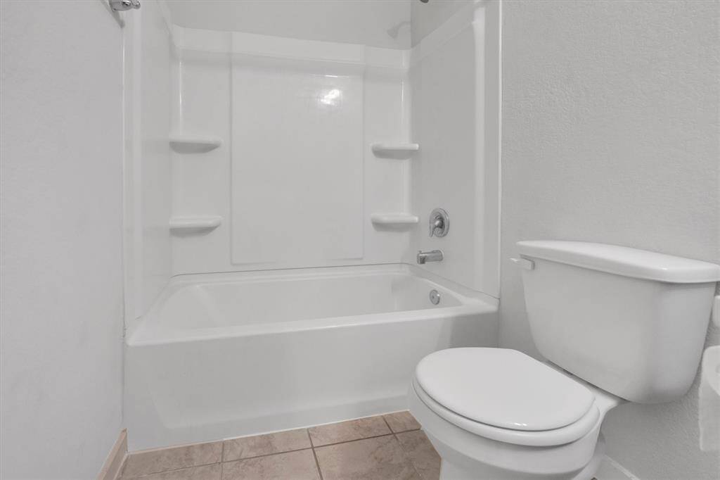 This photo shows a clean and simple secondary bathroom with a bathtub-shower combo and a toilet, featuring neutral tile flooring and white walls.