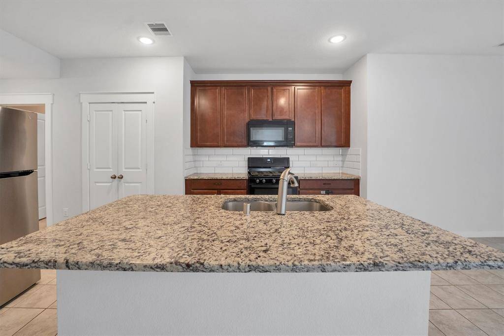 This kitchen features a spacious granite island with a sink, stainless steel appliances, and warm wood cabinetry. The modern look is complemented by a subway tile backsplash and tile flooring.