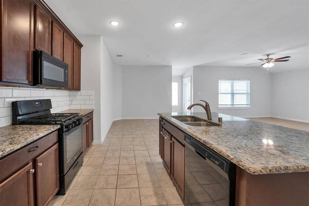 This spacious kitchen features granite countertops, a central island with a sink, and dark wood cabinetry. It opens into a bright living area with tile flooring, recessed lighting, and a ceiling fan. The open layout enhances natural light and flow.