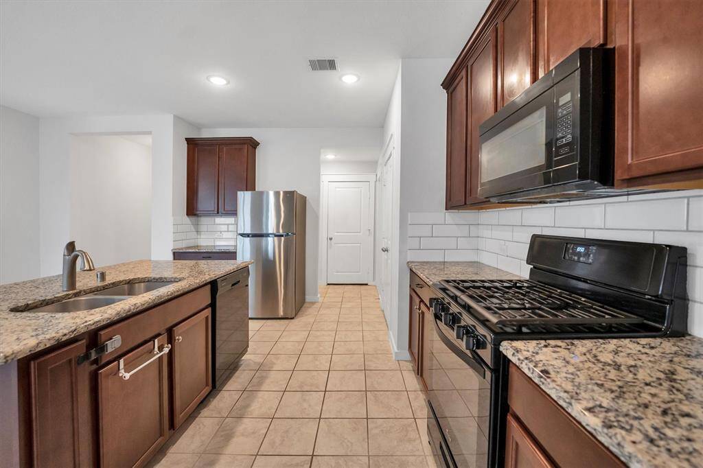 This kitchen features granite countertops, modern stainless steel appliances, and rich wood cabinetry. It offers a spacious layout with a central island, tiled floor, and a subway tile backsplash, combining style and functionality.