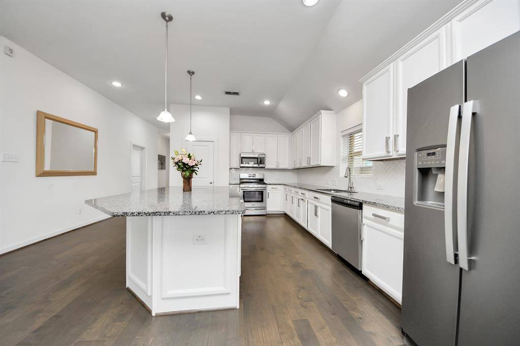 With modern kitchen features sleek white cabinetry, granite countertops, and stainless steel appliances. The spacious island offers ample prep space and room for seating, while pendant lighting adds a stylish touch. The open layout connects to a welcoming hallway with rich floors.