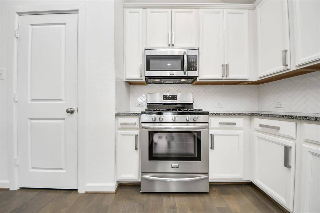 Modern kitchen with sleek stainless steel appliances, including a gas stove and microwave. The space features white cabinetry, granite countertops, and a stylish herringbone tile backsplash, complemented by the floors.