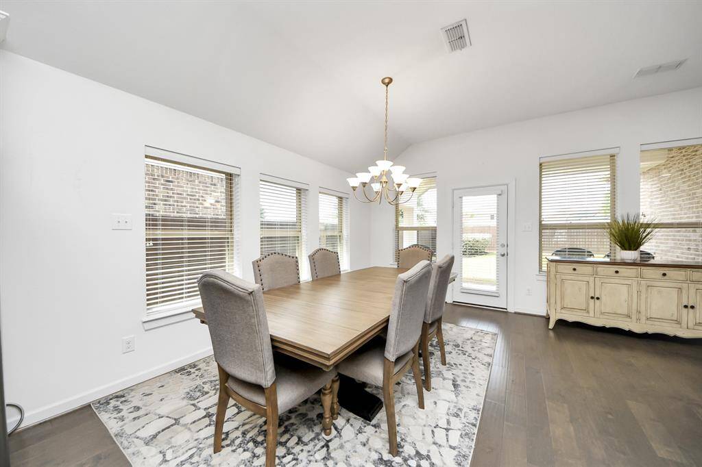 Bright dining area with high ceilings features a large wooden table, elegant chandelier, and ample natural light from multiple windows. The space is enhanced by a patterned rug and a sideboard for storage, creating a welcoming atmosphere for gatherings.