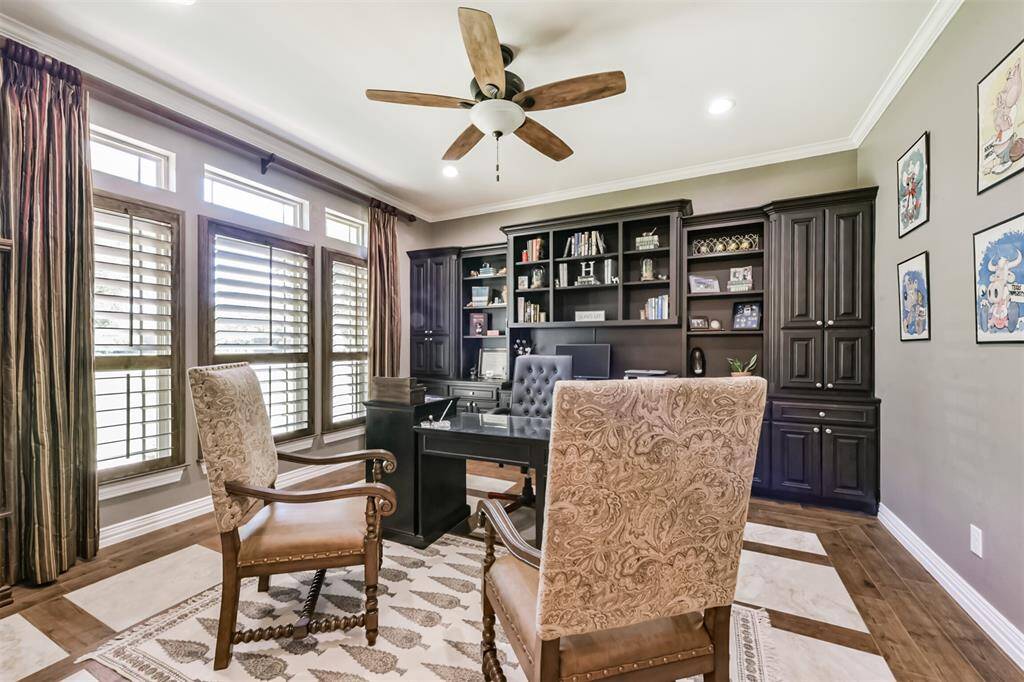 This photo showcases a sophisticated home office with large windows and elegant wooden shutters, providing ample natural light. It features rich wooden cabinetry, a built-in bookshelf and a ceiling fan for comfort.