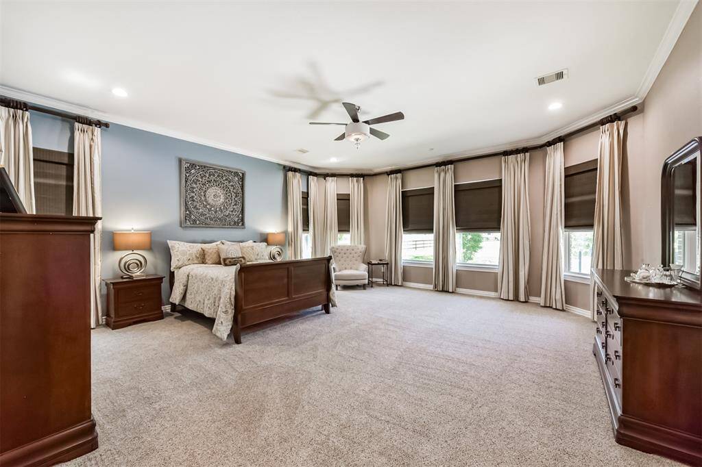 This spacious primary bedroom features neutral tones, large windows with elegant drapes, and plush carpeting. It includes a ceiling fan, ample natural light creating a warm and inviting atmosphere.