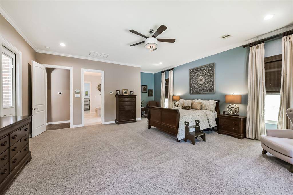 Spacious and inviting primary bedroom with neutral tones, featuring a plush carpet, elegant ceiling fan, and ample natural light through large windows. The room includes  a cozy seating area, and a direct entrance to an en-suite bathroom.