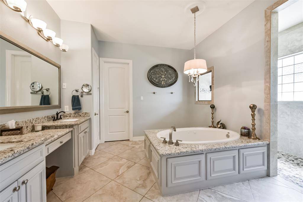 This bathroom features a spacious layout with elegant tile flooring, a large soaking tub with granite surround, and a separate shower. The double vanity offers ample storage and is complemented by stylish lighting fixtures. The soft color palette creates a serene atmosphere.