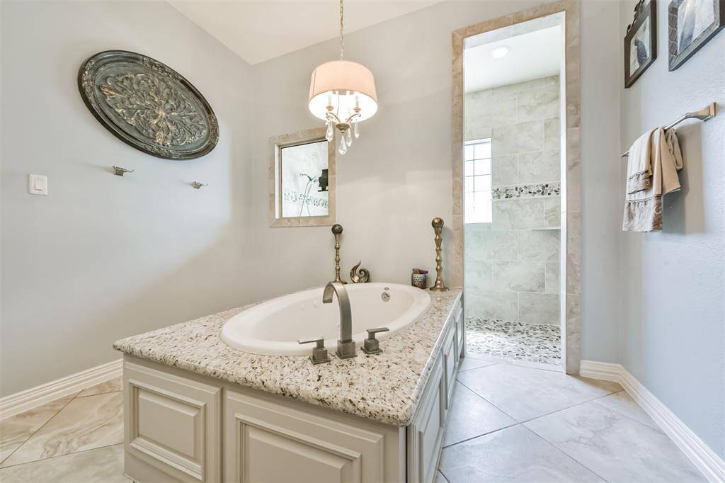 This bathroom features a stylish soaking tub with a granite surround, elegant fixtures, and a large walk-in shower with decorative tile. The neutral palette and soft lighting create a serene atmosphere.