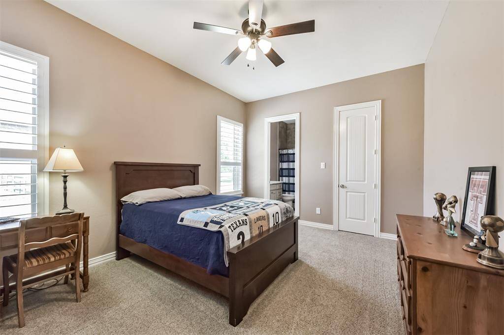 This cozy bedroom features soft beige walls, a ceiling fan, and ample natural light from two windows with shutters.The room has a plush carpet and an en-suite bathroom for added convenience.