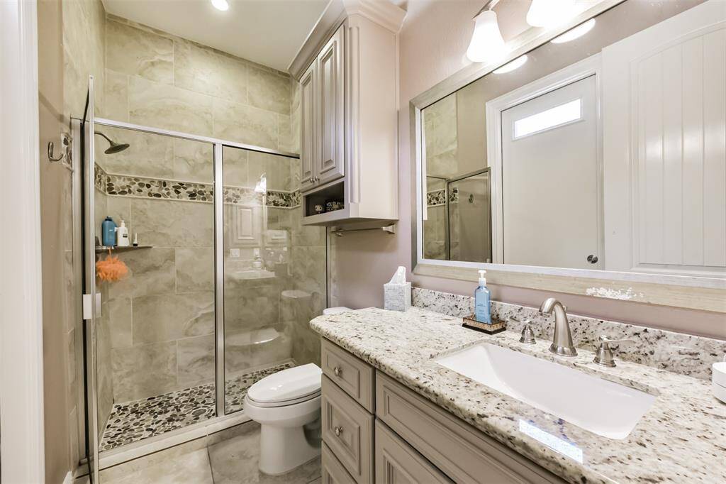 This downstairs full bathroom features a sleek, modern design with a glass-enclosed shower and stone tile detailing. It includes a granite countertop with an under-mount sink, ample cabinet storage, and stylish lighting fixtures.