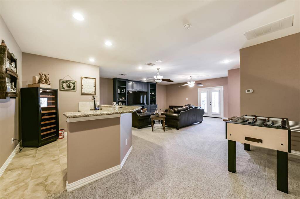 This spacious entertainment room features a cozy seating area, a built-in shelving unit, and large windows for natural light. A corner bar with granite countertops provides many entertainment options. The neutral walls and carpet create a warm, inviting atmosphere.