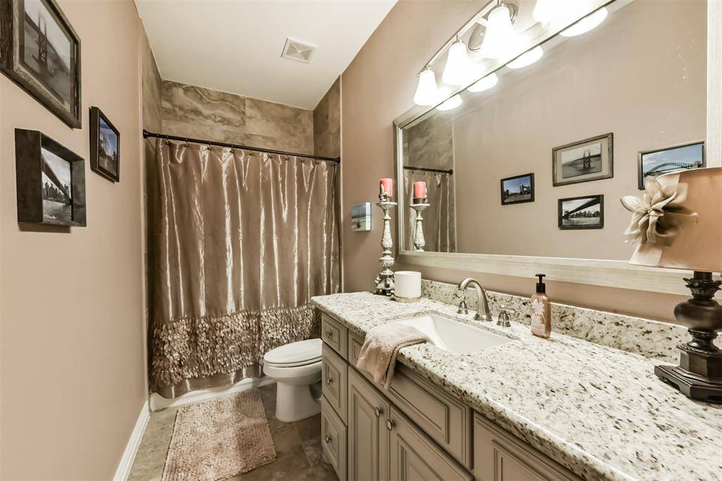 This upstairs bathroom features a granite countertop with a single sink and under-cabinet storage. A large mirror with overhead lighting enhances the space. The shower is enclosed with a decorative curtain. Neutral tones and wall art add a cozy touch.
