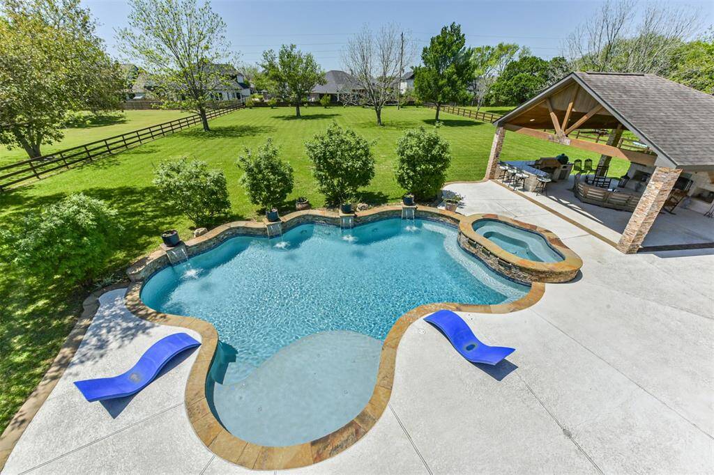 This photo showcases a spacious backyard featuring a uniquely shaped pool with adjacent spa, surrounded by a well-maintained lawn and trees. There's a covered patio area with seating and an outdoor kitchen, perfect for entertainment and relaxation.