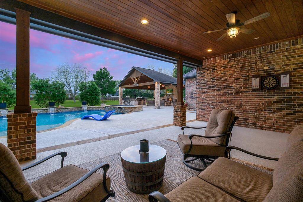 This photo features a spacious covered patio with comfortable seating, a ceiling fan, and a view of a backyard pool. The pool area includes a slide and a covered outdoor kitchen, perfect for entertaining. The setting is enhanced by a scenic, lush landscape and a beautiful sky.