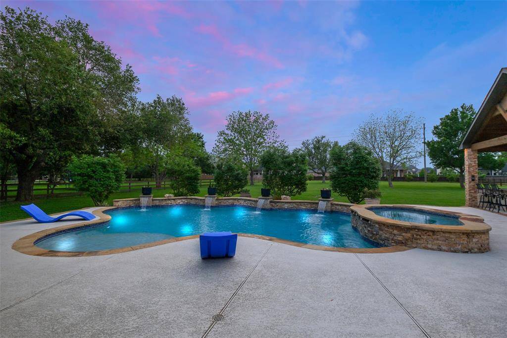 This photo showcases a serene backyard with a beautifully lit pool and spa, surrounded by lush greenery and ample patio space, perfect for relaxation and entertaining.