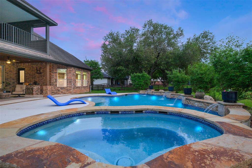 This inviting backyard features a spacious pool and spa with elegant stonework, surrounded by lush greenery. The brick exterior of the house complements the serene setting, and the patio offers a perfect spot for relaxation.