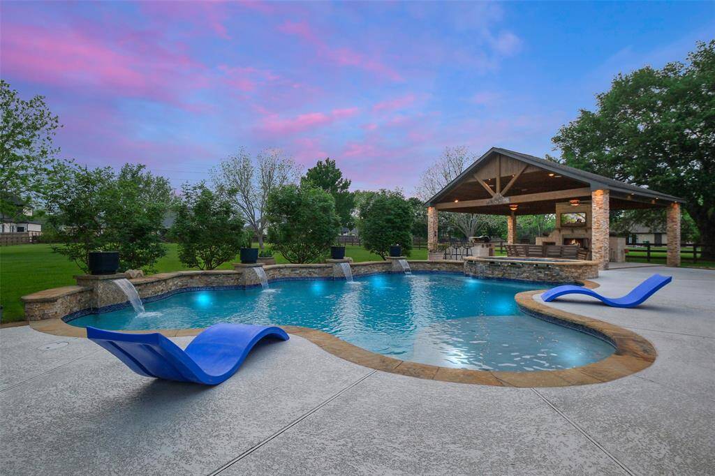 This photo showcases this stunning backyard with a custom-shaped pool featuring built-in water features, and a spacious covered patio area perfect for entertaining, all set against a backdrop of lush greenery and a vibrant sunset sky.