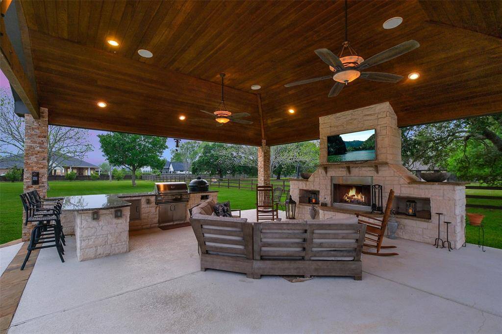 Luxury, spacious outdoor living area with a wooden ceiling, featuring a cozy fireplace and mounted TV. It includes a built-in grill and bar seating, set in a lush, landscaped backyard, perfect for entertaining or relaxing.
