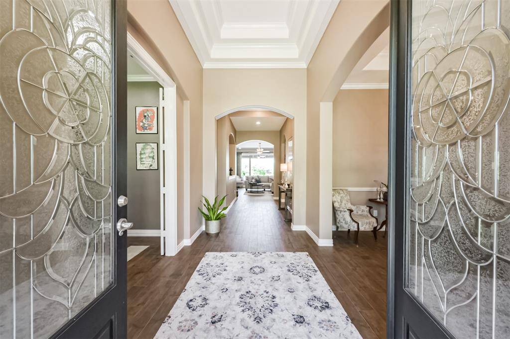 This welcoming entryway features elegant glass doors and a wide hallway with warm wood tile flooring. It leads to a bright and inviting living area, with stylish arches and decorative moldings adding charm and sophistication.