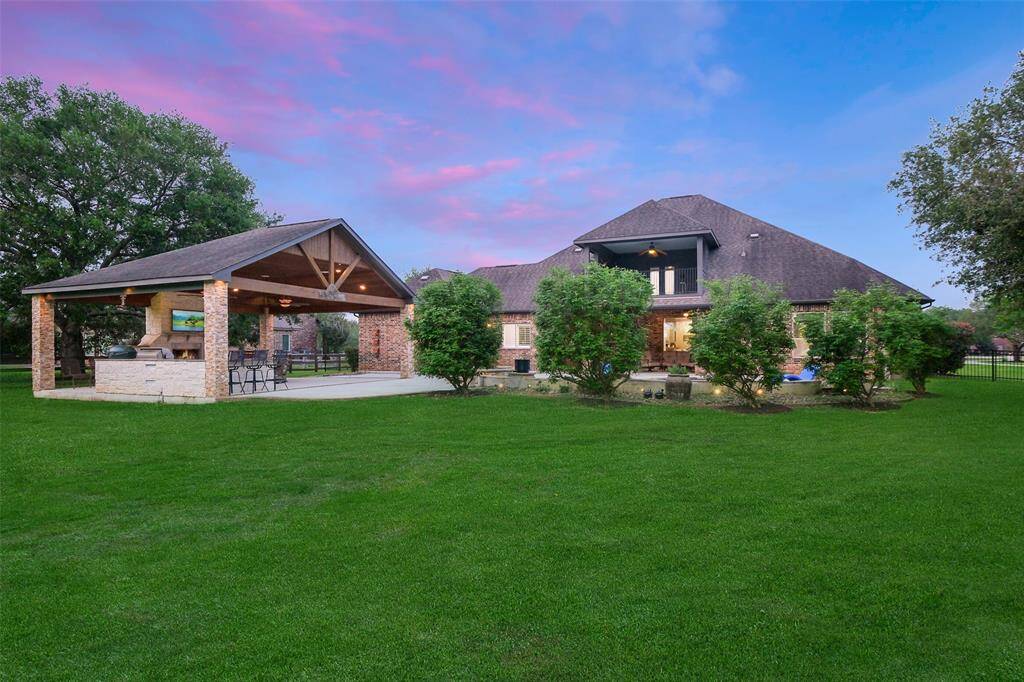This photo showcases a spacious backyard with a well-maintained lawn, a large house featuring multiple rooflines, and a covered outdoor dining area with built-in amenities. The serene setting is enhanced by surrounding trees and a vibrant sky.