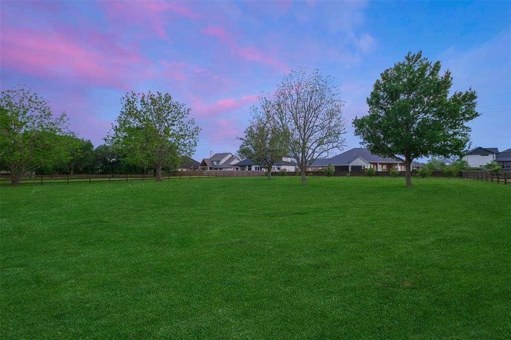 This photo showcases a spacious, lush green yard with a few mature trees, enclosed by a fence, and neighboring houses in the background under a vibrant, colorful sky. Ideal for those seeking a serene, open outdoor space.