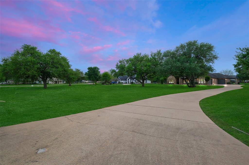 This photo showcases a spacious property with a long, curved driveway leading to a house surrounded by lush green lawns and mature trees, set against a vibrant sunset sky.