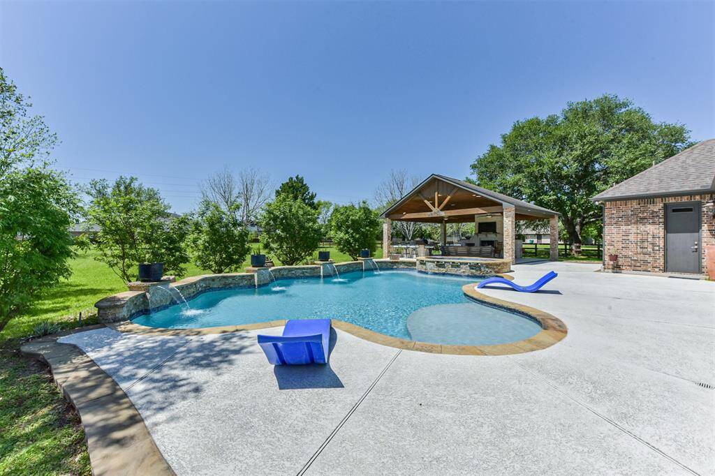 Spacious backyard featuring a modern in-ground pool with a fountain, surrounded by greenery. Includes a covered patio area with ceiling fans and outdoor kitchen, perfect for entertaining.