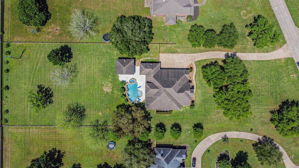 Aerial view of a spacious property featuring a large home with a swimming pool, surrounded by extensive green lawns and mature trees. The property is located on a quiet street with a curved driveway and ample space for outdoor activities.