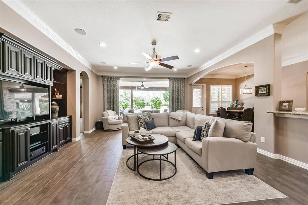 Spacious living room with an open floor plan, featuring a large sectional sofa, elegant built-in entertainment center, and ample natural light from large windows. The room seamlessly connects to the dining area, creating a welcoming space for relaxation and entertaining.