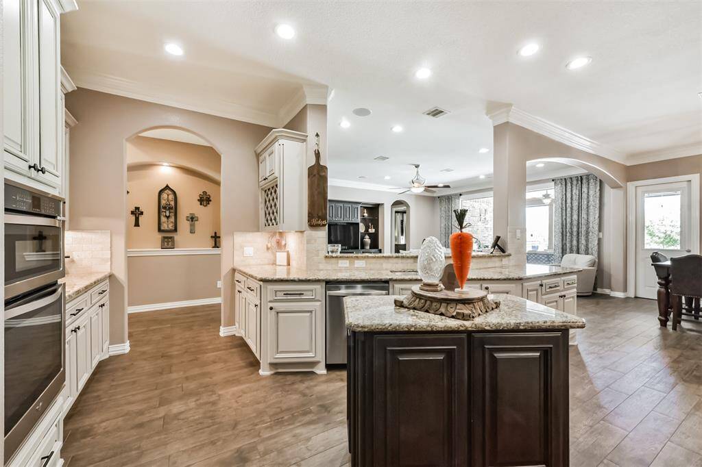This open-concept kitchen features elegant cabinetry, granite countertops, and a central island with decorative accents. It seamlessly connects to a spacious living area with a ceiling fan and large windows, offering a bright and inviting atmosphere.
