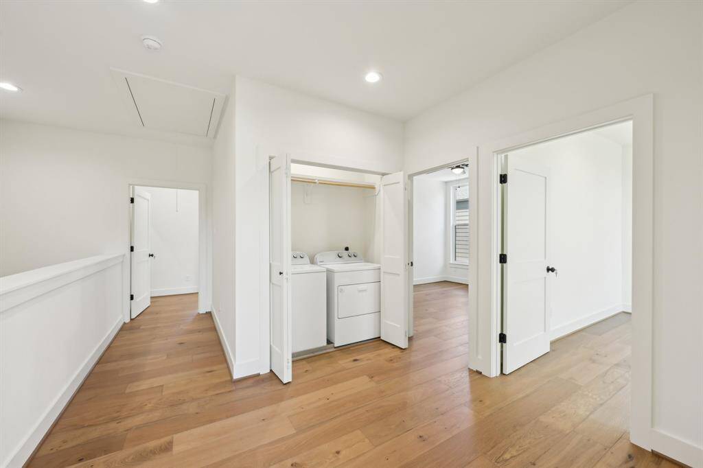 This photo shows a bright, open hallway with hardwood floors, featuring a compact laundry area with washer and dryer behind double doors.