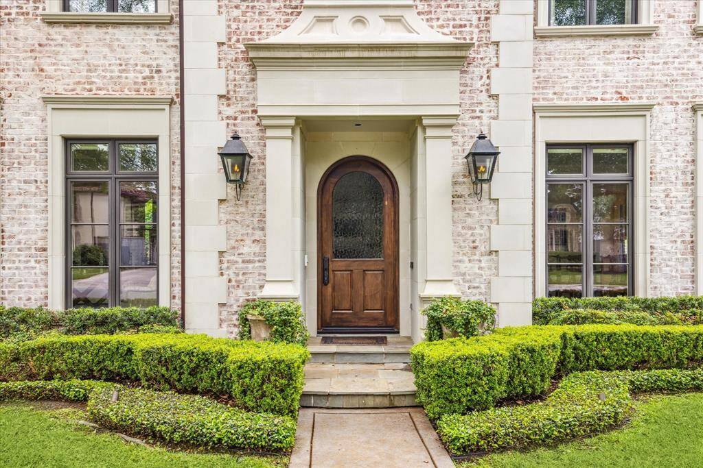 A warm welcome begins at this stunning arched mahogany front door, framed by timeless cast-stone trim and flanked by elegant gas lanterns. The covered entry showcases refined architectural detailing and opens to a lushly landscaped path bordered by sculpted boxwoods.