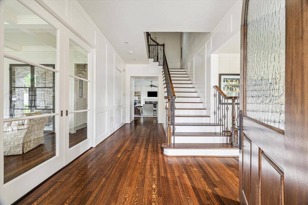 Step inside to a light-filled foyer with rich hardwoods, crisp panel molding, and soaring ceilings that create an immediate sense of elegance. A sweeping staircase with wrought iron balusters anchors the space, while glass-paneled doors offer a glimpse into the office area, setting the tone for the home’s thoughtful design and timeless sophistication.