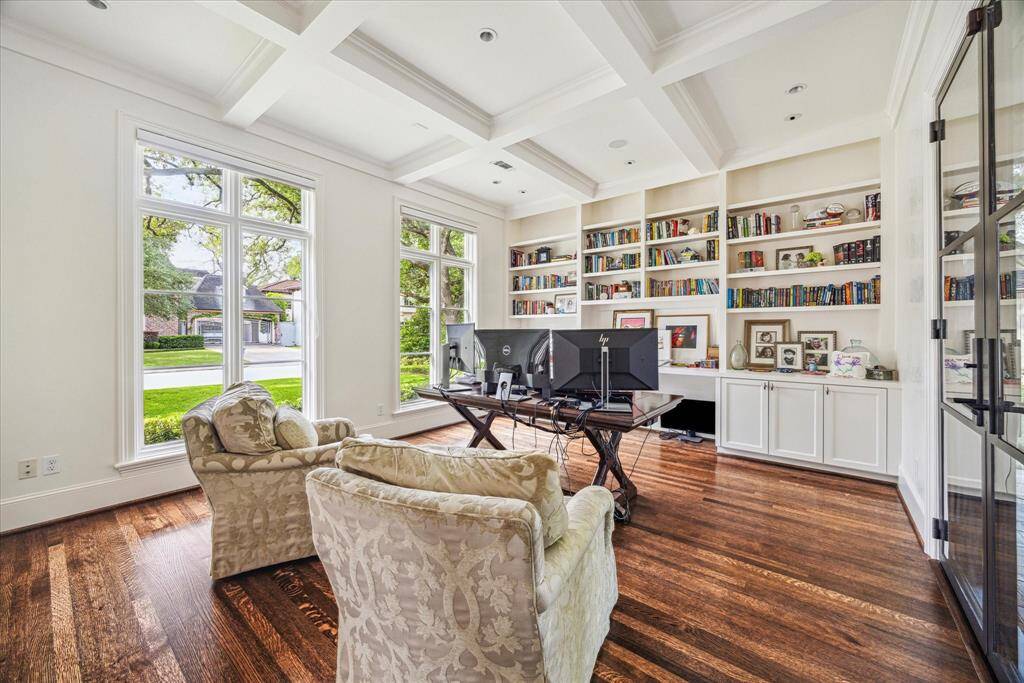 This stunning home office offers a serene and sophisticated workspace with floor-to-ceiling built-ins, hardwood floors, and a coffered ceiling that adds architectural depth. Sunlight pours through oversized windows with views of the front lawn, while steel-framed glass doors provide seamless access to the backyard and patio area with tranquil pool views.