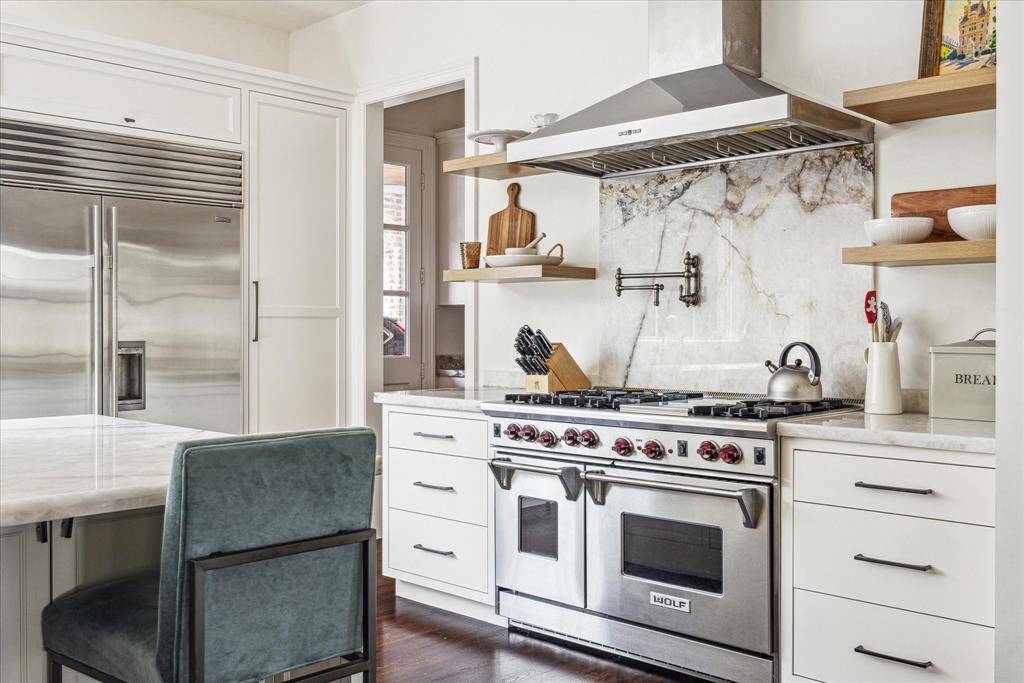 The chef's kitchen combines high-end style with everyday functionality, featuring premium appliances including a Wolf oven range, a built-in Sub-Zero refrigerator, a statement quartzite backsplash, a convenient pot filler,  and open shelving for a clean, modern aesthetic.