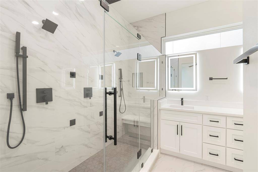 Modern bathroom with dual wall shower heads, body sprays, and rainfall shower head, and a sleek vanity with ample storage and backlit mirrors.
