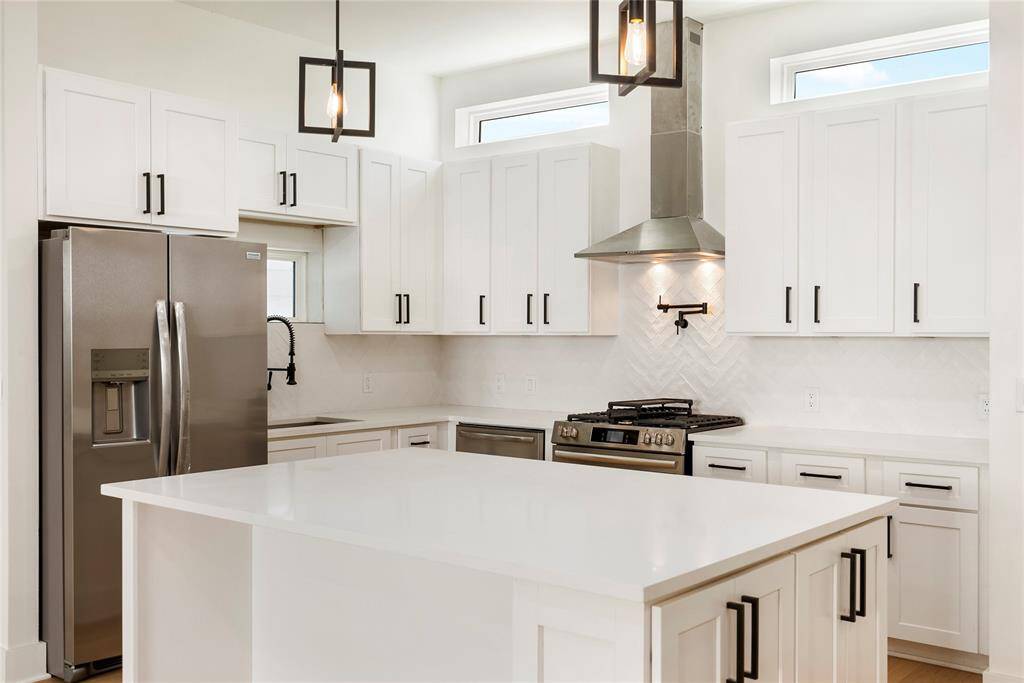 This is a modern kitchen featuring white cabinetry, stainless steel appliances, a gas stove with pot filler, and a central island with a white countertop. It has a clean, contemporary design with a herringbone tile backsplash.