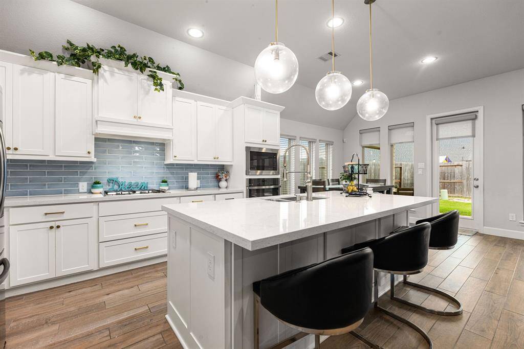 This stunning kitchen displays such a beautiful contrast with the white cabinets, neutral paint, quartz countertops, blue modern backsplash, SS appliances, oversized kitchen island with extra storage, breakfast bar, recessed lighting, high ceilings and modern pendants over the bar.