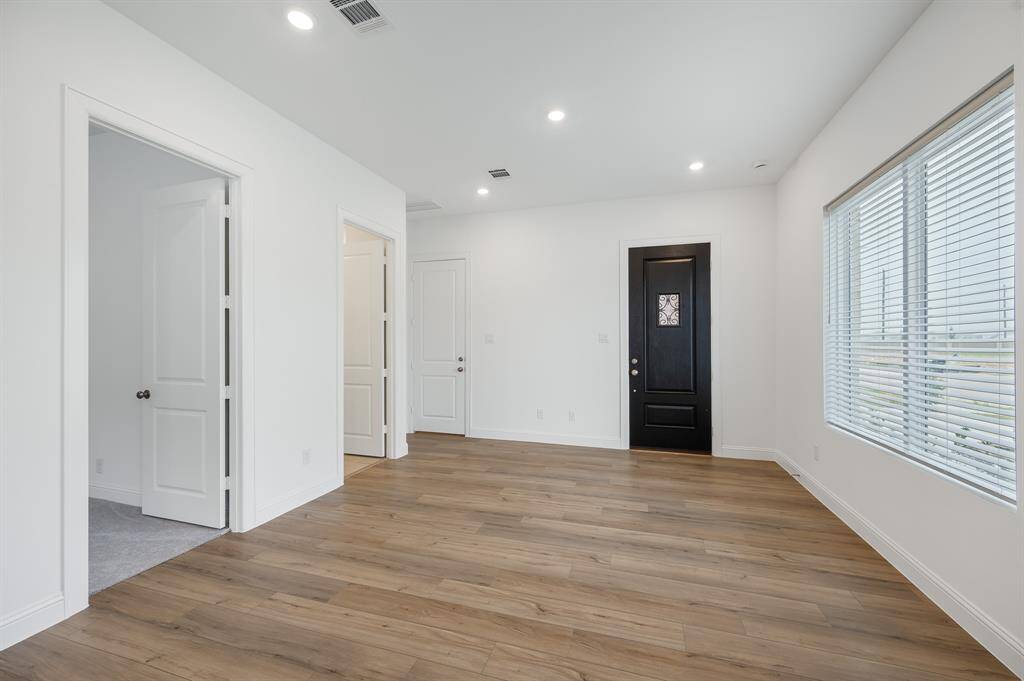 Next Gen® Suit offers bright living room with wood look flooring, recessed lighting, and a large window with blinds. Features multiple white doors, including a stylish black front door with decorative glass.