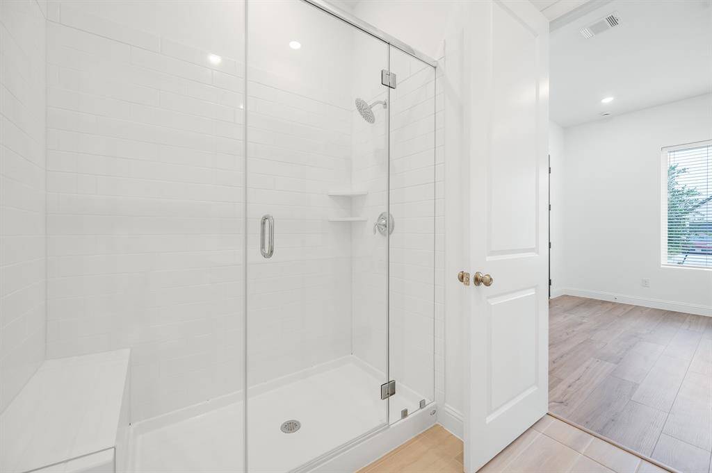Next Gen® Suite modern bathroom with a glass-enclosed shower featuring white tile. The room has wood-style flooring and an adjacent living area with natural light from a window, creating a clean and inviting atmosphere.