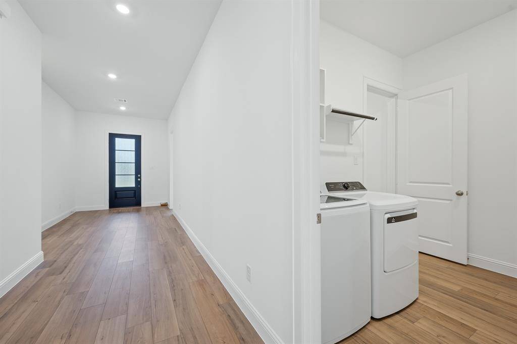 This photo shows a bright hallway with wood flooring, recessed lighting, and a modern black door. To the right, there's a laundry area with a washer and dryer, and shelving above.