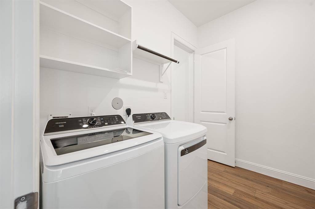 This is a bright, modern laundry room featuring a washer and dryer, open shelving, and a hanging rod, all set against clean white walls and wood-style flooring.