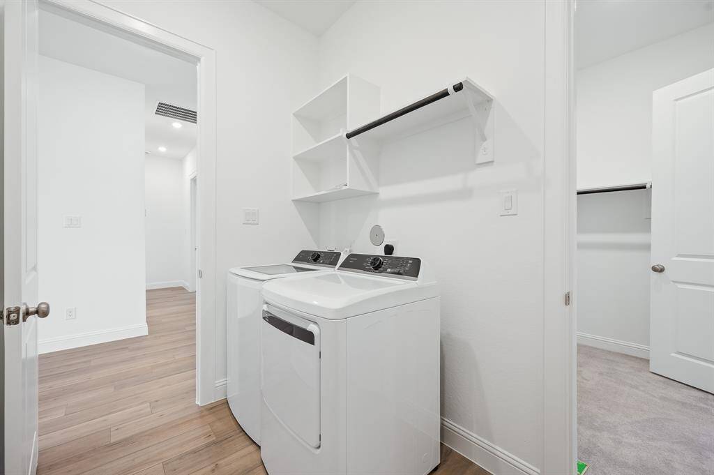 This photo shows a bright, modern laundry room with a washer and dryer, white walls, and light wood flooring. It includes shelving and hanging space for convenience, and connects to other rooms via white doors.