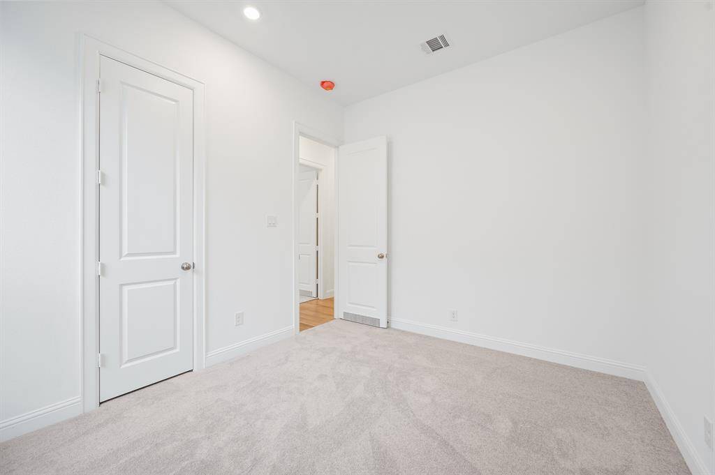 Secondary bedroom with white walls and a light beige carpet. It features a closed closet door and an open door leading to a hallway with wooden flooring. Recessed lighting is visible on the ceiling.