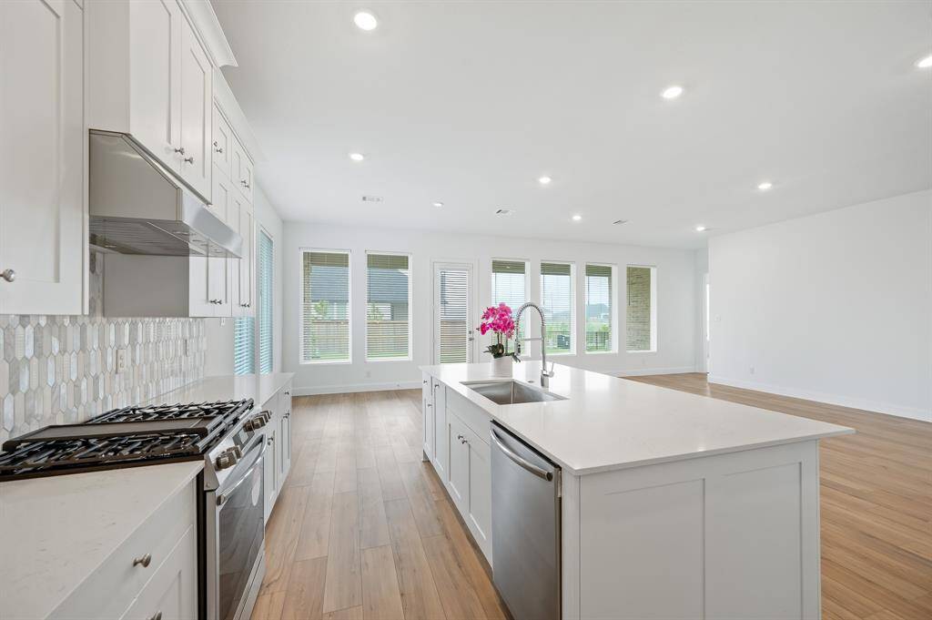Bright, modern kitchen with white cabinetry, stainless steel appliances, and a large island with sink. Open layout, wood style flooring, and ample natural light from multiple windows.