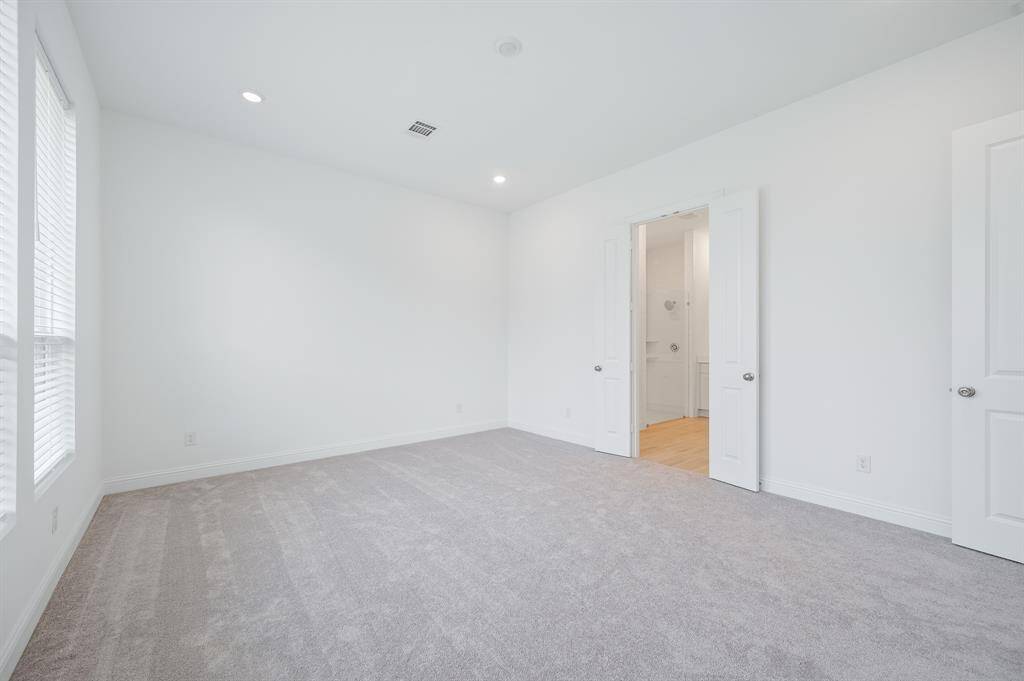 Bright, spacious primary room with neutral-color carpeting and white walls. Features include recessed lighting, large windows for natural light, and an open doorway leading to an adjoining room.