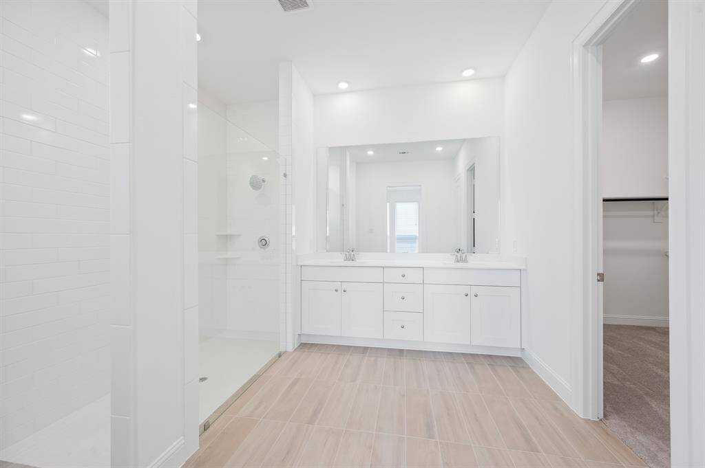 A spacious, modern primary bathroom suite featuring a double vanity with ample storage, a large mirror, and a glass-enclosed shower. The room is bright with recessed lighting and a neutral color palette, creating a clean and inviting atmosphere.