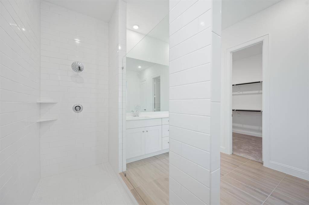 A modern, bright  primary bathroom with a spacious walk-in shower featuring white subway tiles. There's a large mirror above a sleek vanity, and an adjacent walk-in closet with ample shelving.